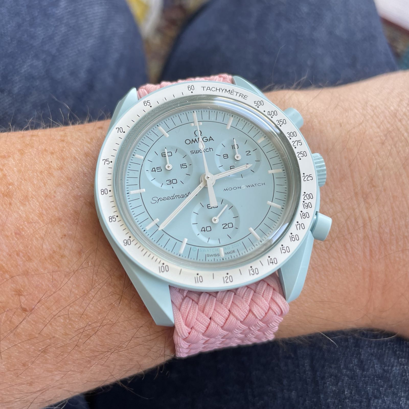 A baby blue watch on a pink perlon strap. The watch case and dial are baby blue, while the tachymeter bezel is white. The watch has three sub registers at 2:00, 6:00, and 10:00.