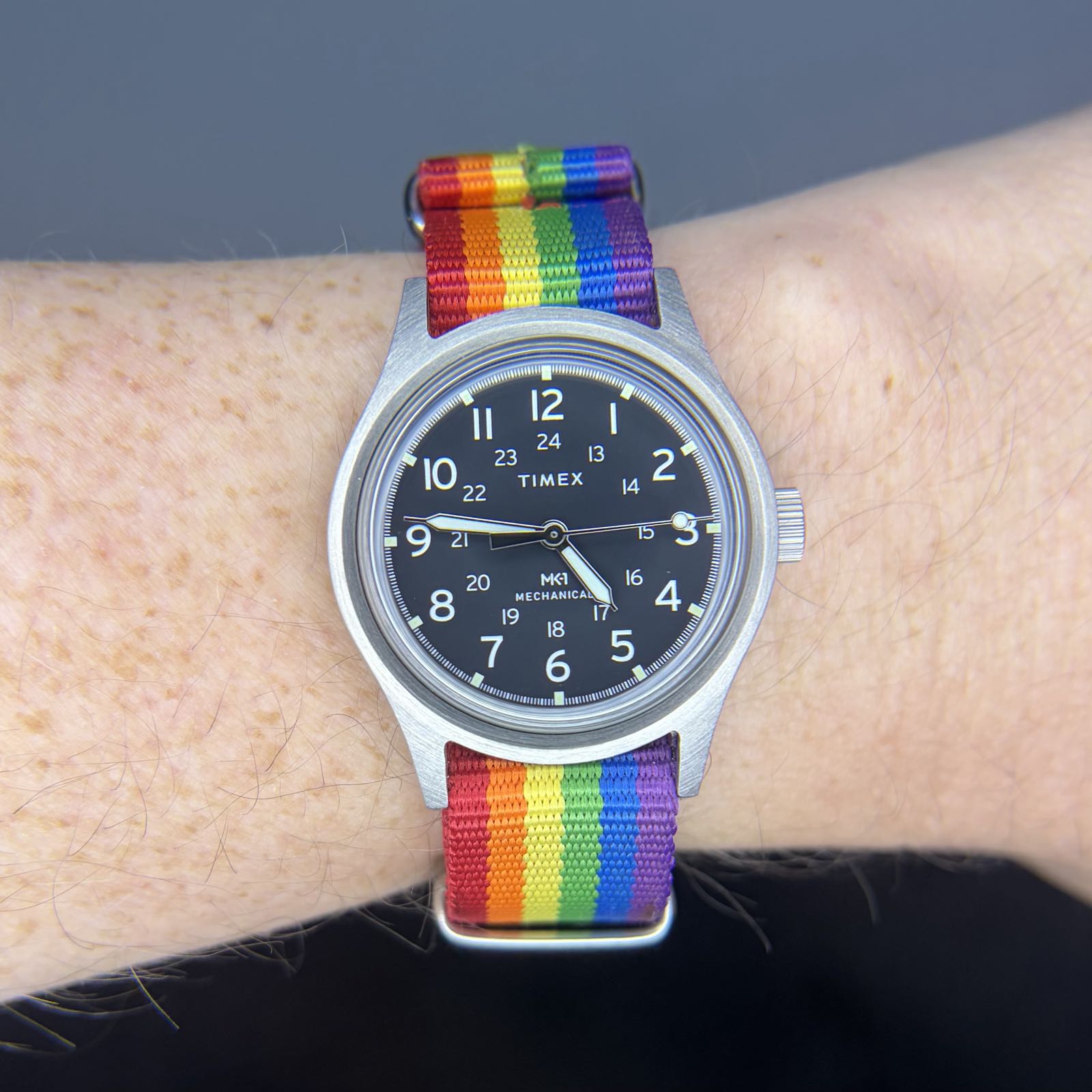 A simple watch is on a Pride flag strap. The dial is black, with simple white arabics at the hours.