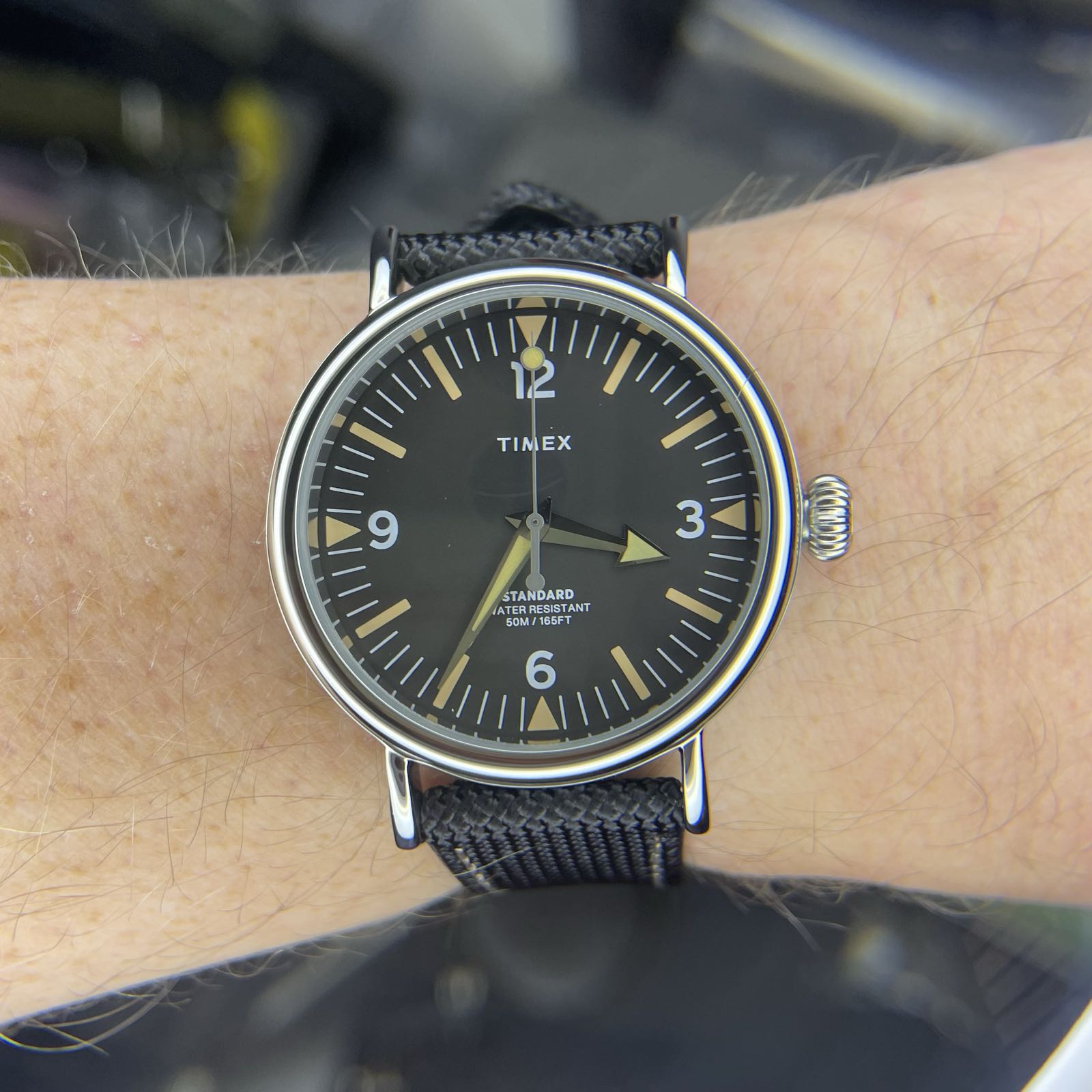 A simple field watch on a black fabric strap. The face is dark gray, with faux patina hour markers. White arabics are in 12–3–6–9 formation, and the hour hand has a broad arrow tip.