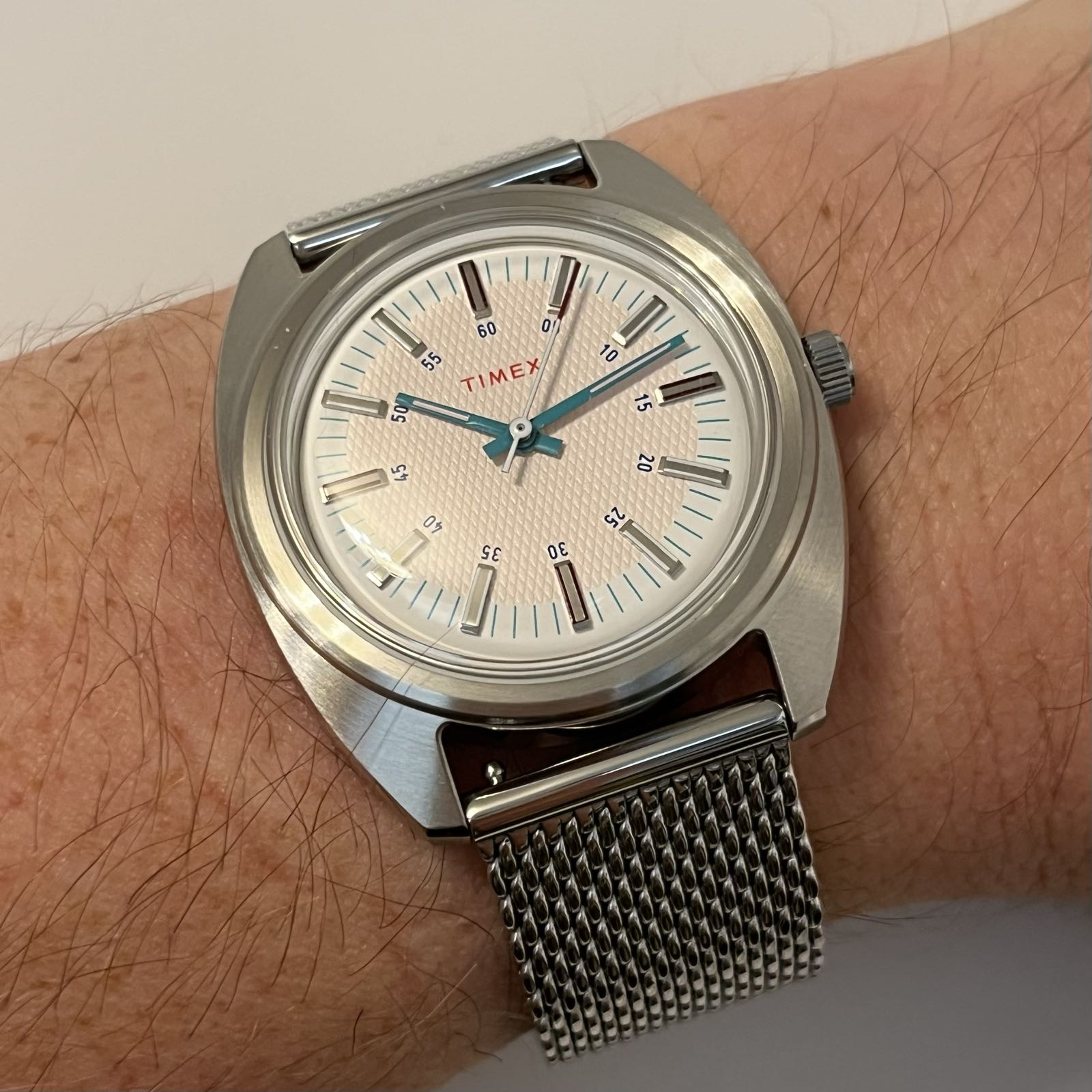 A cushion-case watch is on a mesh bracelet. The dial of the watch is a subtle sunburst, a bit pink in the middle, fading to white at the edge, with a honeycomb pattern on it. The hands are green.